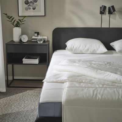 Bedroom scene with SANDGRÄSMAL duvet on bed, gray nightstand holding books, clock, and plant; black headboard, wall lamps.