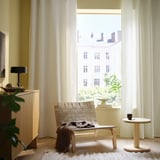 Modern living room with large window, sheer SANELA white curtains, light wood furniture, and a cozy woven chair with a blanket.