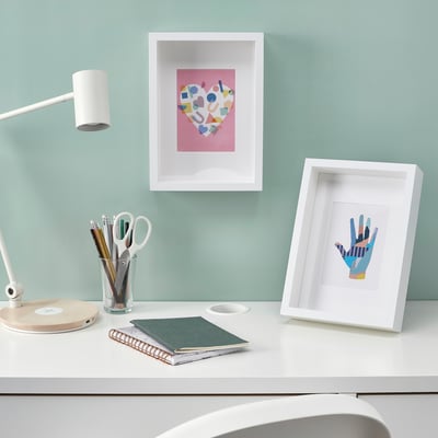 White desk, blue wall, lamp, notebooks, 2 frames