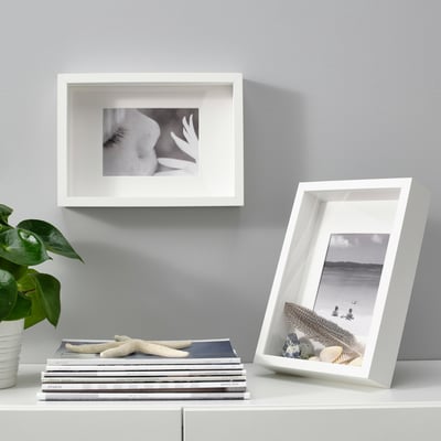 Two SANNAHED frames: one on wall, one on shelf with books, holding flower and beach photos; plant beside books.
