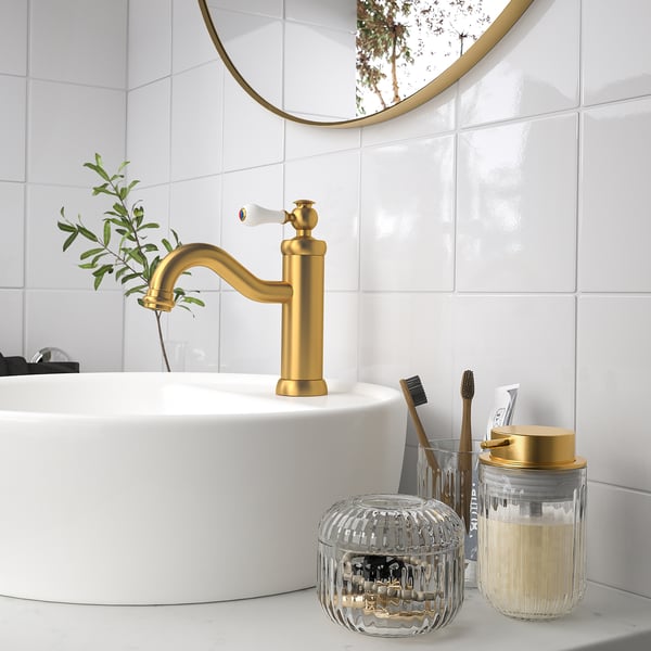 Gold faucet, white sink, round mirror, toiletries, SILVTJÄRN soap dispenser. Glass adds luxury.