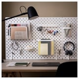SKÅDIS white pegboard with accessories: lamp, scissors, headphones, mesh organizers. Organized home office setup.