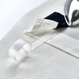 A shiny silver iron rests on white SLÅNHÖSTMAL curtain, showcasing its sleek eyelets for easy hanging.
