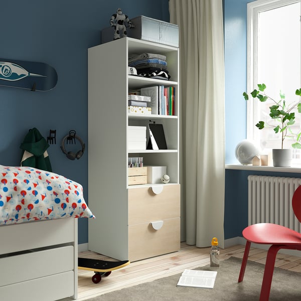 Modern blue bedroom with white bookcase holding items, red chair, colorful bed pillows.