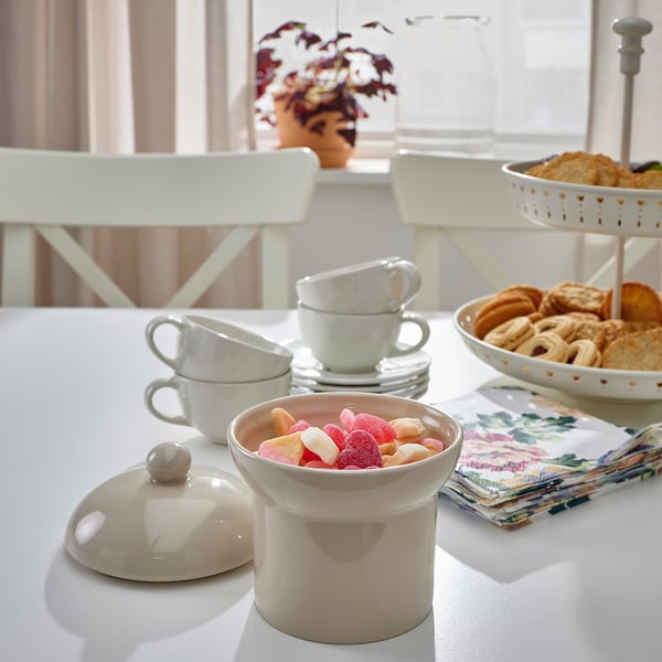 A kitchen counter displays a white ceramic jar filled with colorful candies, next to stacked white cups and a two-tiered tray of pastries.