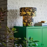 Forest-themed table lamp STOCKHOLM 2025 on green cabinet, brick wall background, surrounded by small plants.