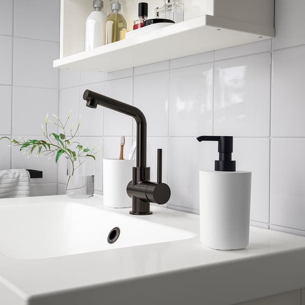 Modern bathroom sink with STORAVAN black faucet and white accessories. Pump dispenser, toothbrush holder, and tray on clean white countertop.