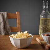 Ceramic bowl of chips on wooden table with oil and nuts, chair in back.
