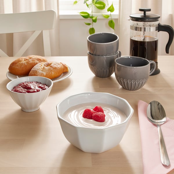 Wooden table set with coffee, pastries, and white ceramic bowls and cups. Bowl in front has yogurt topped with raspberries.