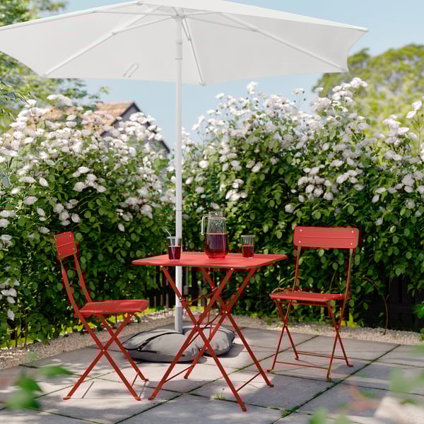 Bright red metal patio set with white umbrella, two folding chairs, and square table under blooming bushes, perfect for outdoor relaxation.