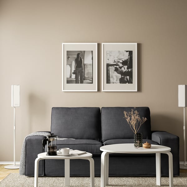 Minimalist living room: gray sofa, two black & white framed photos, round white coffee tables, SYMFONISK floor speakers.