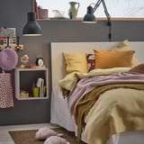 Bedroom with cozy bed, mustard bedding, wall shelf with books, lamp, clock, and decorative items.