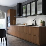Modern kitchen with wooden cabinets, black handles, marble backsplash, and black appliances. Natural light illuminates a white pendant lamp.