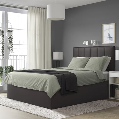 Modern dark gray TJÅLLE bed with green linens, large against blue wall. It has a padded headboard and bedside lamps. Rug and balcony seen.