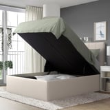 Modern bedroom with TJÅLLE ottoman bed, white headboard, green blanket, and plant. Open storage in bed, nightstand, and soft lighting.
