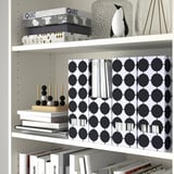 White bookshelf with TJENA magazine holders, board games, and decorative items. The holders organize papers and magazines with a stylish pattern.