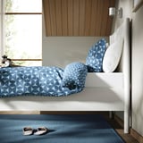 Bed with patterned blue bedding, slatted headboard, wall lamp, rug, and slippers, offering cleanliness and storage.