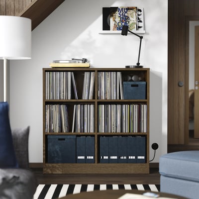 A TONSTAD wooden shelf unit with vinyl records and storage boxes. Its brown, rectangular, and sits between furniture, with a lamp and chair nearby.
