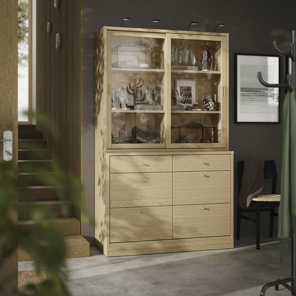 A modern wooden cabinet named TONSTAD in oak finish with glass doors, displaying decorative items, and six drawers for storage.