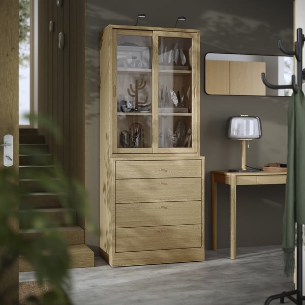 Wooden light brown cabinet with glass doors and drawers. Displays decorative items and organizes contents. Sleek design for modern spaces.