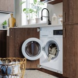 Modern kitchen with integrated white washing machine and dryer, wooden cabinets, and a wicker laundry basket.