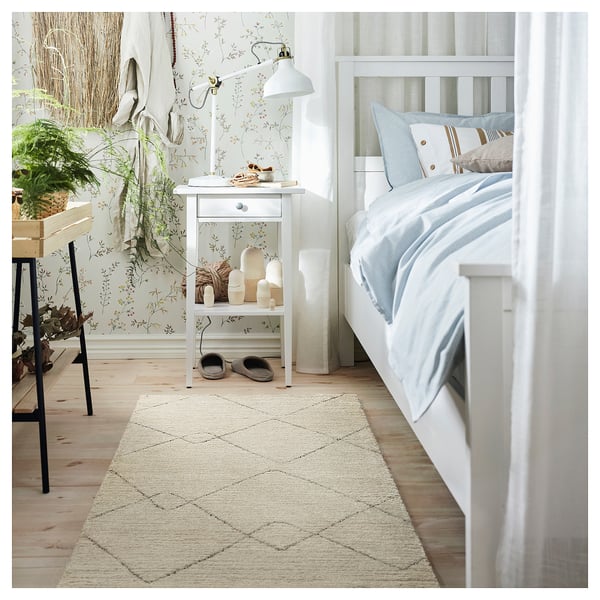 Cozy bedroom with white bed, small table, lamp, and a beige TVERSTED rug. Decor includes wallpaper, curtains, and plant.