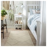 Cozy bedroom with white bed, small table, lamp, and a beige TVERSTED rug. Decor includes wallpaper, curtains, and plant.