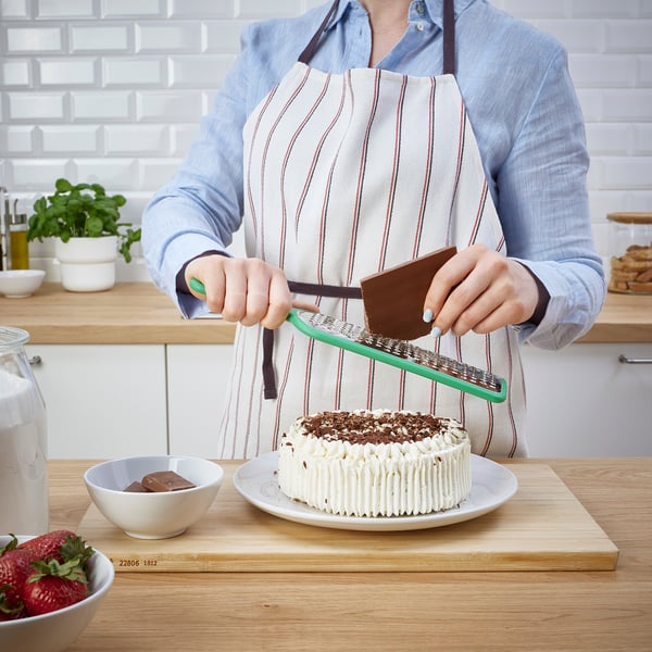 Person grates chocolate on a green grater over a cake with white frosting. The grater is held over a wooden board with a bowl of strawberries nearby.