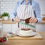 Person grates chocolate on a green grater over a cake with white frosting. The grater is held over a wooden board with a bowl of strawberries nearby.