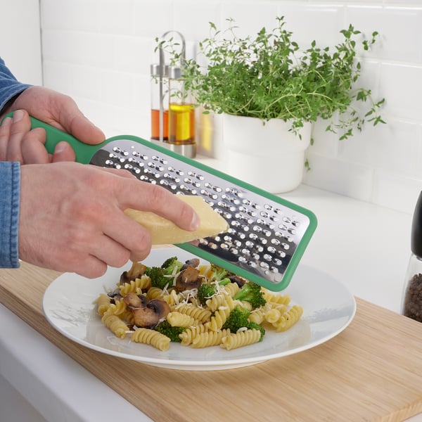 A person grates cheese over pasta with broccoli using a green UPPFYLLD grater, efficiently enhancing the dishs flavor.