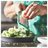 A person grates cheese onto broccoli with a green UPPFYLLD grater.