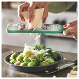 Hands grate cheese with green UPPFYLLD grater over a bowl of broccoli and brussels sprouts on a countertop.