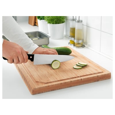 A person slices cucumbers using a large, sharp kitchen knife on a wooden cutting board.