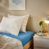 Cozy bedroom with blue VÅRVIAL bedding, white pillows, nightstand, lamp, plant, and blue lotus art.