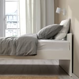 Modern white bed frame with grey bedding, wall lamp, and curtains.