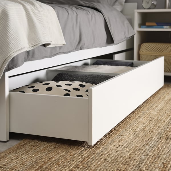 A white bed frame with an open bottom, showing a narrow storage drawer with polka dot fabric partially pulled out, offering under-bed storage.