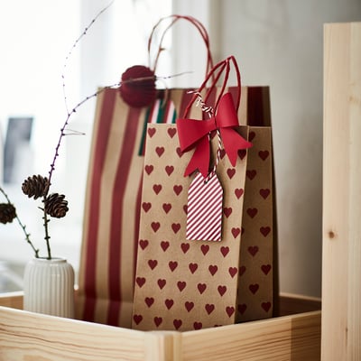 Two gift bags with heart patterns and red bows, one large and one medium, placed in a wooden crate with a small vase containing dried flowers.
