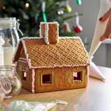 A gingerbread house being decorated with white icing. The house has a brown color, waffle-textured roof, and a chimney with white decorative lines.