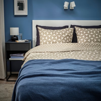 Mesa de cabeceira muito menor com lâmpada ao lado de cama grande, roupa de cama geométrica