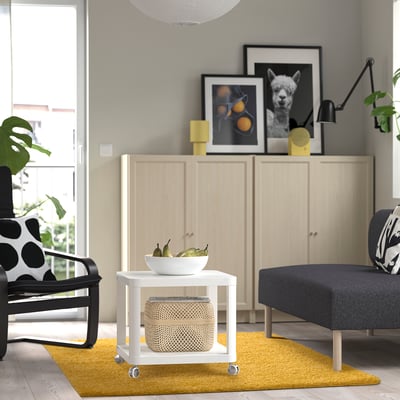 Sala de estar moderna com tapete amarelo, armário de madeira, cadeira preta, sofá cinzento e mesa lateral branca.