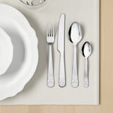 Quatro utensílios de prata polida - garfo, faca, duas colheres - colocados em um jogo americano branco ao lado de uma tigela branca. Ideal para refeições elegantes.