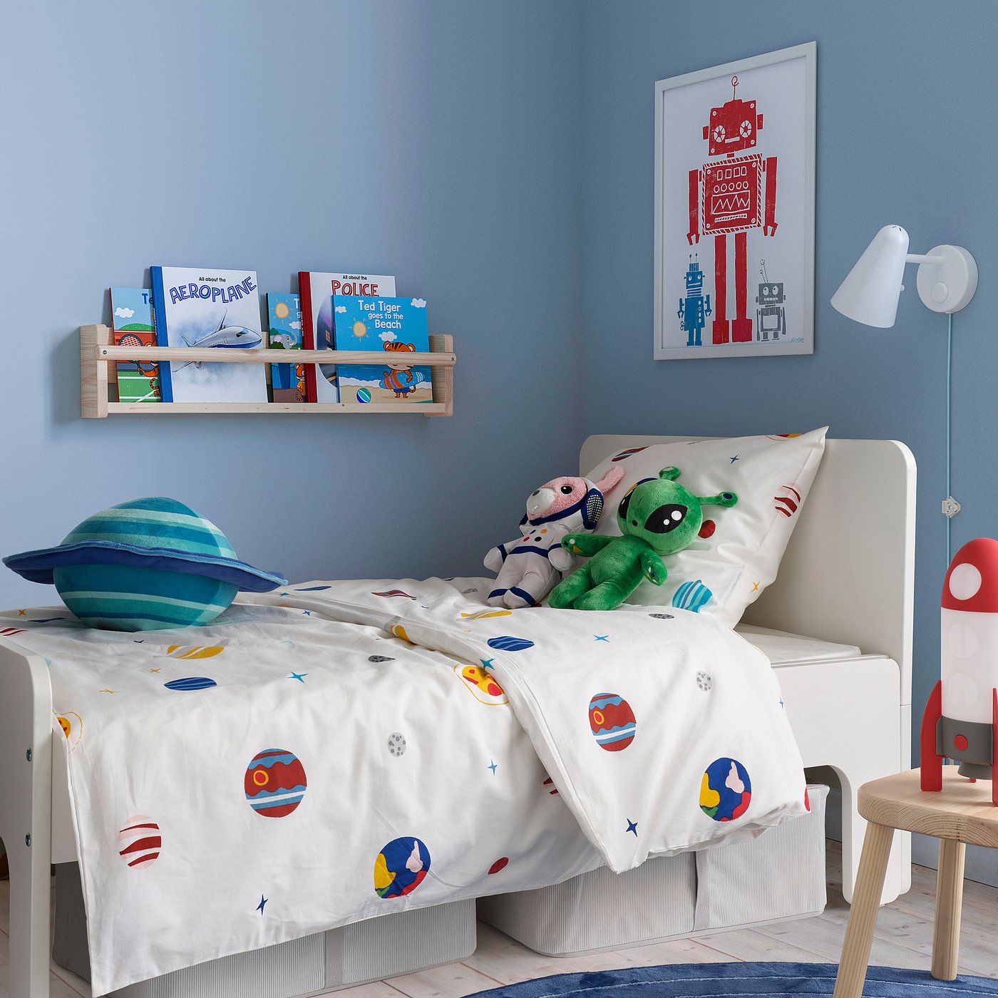 Quarto infantil com edredom AFTONSPARV, planetas, estrelas, parede azul, estante, pôster de robô vermelho, travesseiro planeta azul, brinquedo verde, lâmpada de foguete vermelha.