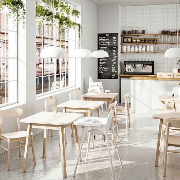 Café minimalista com mesas brancas, cadeiras e grandes janelas. Plantas suspensas e placa de menu. Design luminoso e espaçoso.