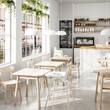 Café minimalista com mesas brancas, cadeiras e grandes janelas. Plantas suspensas e placa de menu. Design luminoso e espaçoso.