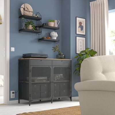 Prateleira de metal preto com plantas, decoração e eletrônicos, cabendo no canto da sala ao lado da poltrona bege.