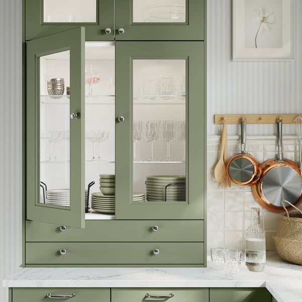 Cozinha moderna com armários AXSTAD verdes, portas de vidro com pratos, bancada em mármore, utensílios suspensos e artigos decorativos.