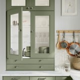 Cozinha moderna com armários AXSTAD verdes, portas de vidro com pratos, bancada em mármore, utensílios suspensos e artigos decorativos.