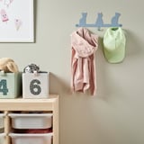 Espaço infantil com cabides em forma de gato segurando um moletom rosa e boné de beisebol verde; abaixo, caixas de armazenamento com brinquedos e bichos de pelúcia.