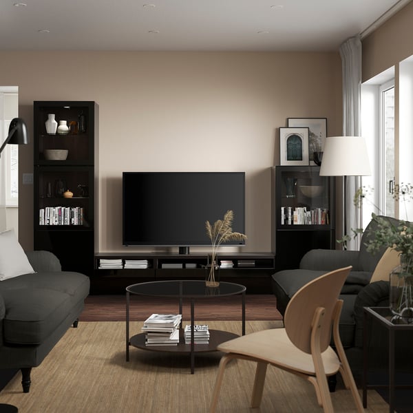 Sala de estar moderna com móvel de TV BESTÅ, sofá cinza, cadeira, tapete e mesa. TV ladeada por prateleiras com livros e decoração.