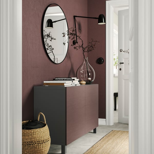 Entrada moderna com aparador BESTÅ em roxo escuro e preto. Inclui espelho, lâmpadas e cesta. Pavimento cinzento claro e porta branca.